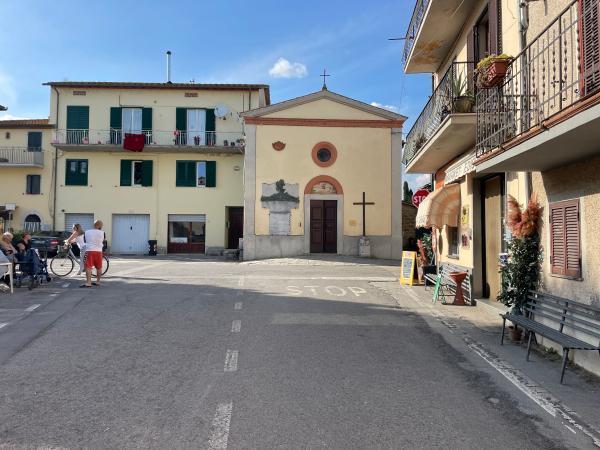 Strada che arriva nella Piazza di Borghetto. Chiesa beige, con portale rosso e croce in legno sul lato destro. A sinistra, alcune case.